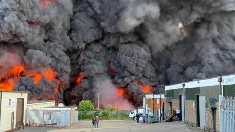 Huge cloud of dark grey smoke billowing from the ground, with flame visible in the lower half. There are bushes in front of the smoke. There is a single-storey building to the right and another to the left. There are people standing on the yard in between.
