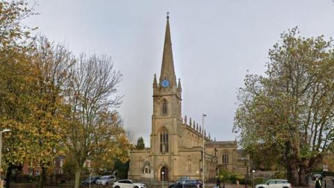 Honey-coloured stone church with spire and blue clock flanked by trees