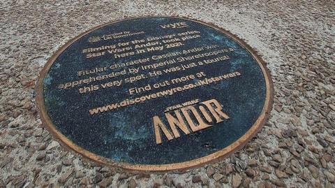 Distance image of the plaque on the promenade showing the cafe in the background and large sandstone wave sculptures to the left