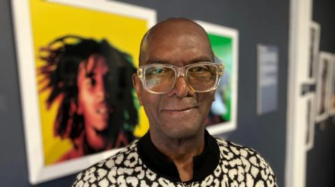 A close-up portrait shows a man standing in a gallery space, wearing large transparent-framed glasses and a patterned black-and-white jacket. Behind the figure, slightly out of focus, are brightly coloured framed photographic artworks, including a vivid yellow and green portrait of Bob Marley.