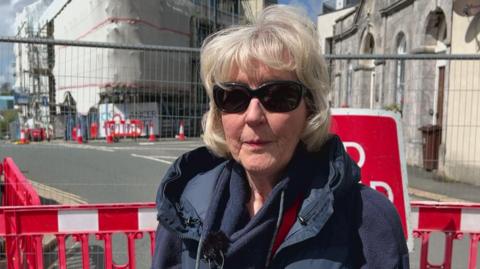 The image shows a person standing in the foreground behind red and white temporary road barriers. The person is wearing a dark padded gilet over a long‑sleeved top and has a red lanyard around their neck. A small microphone is clipped to their clothing, suggesting they are being interviewed on camera.