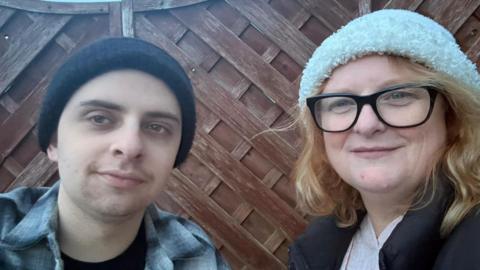 Noah Herniman ar y chwith yn gwisgo het due a chrys llwyd. Shelley Herniman ar y dde yn gwisgo het gwyn a sbectol.