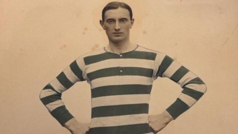 A footballer pictured in the early 20th century. His football shirt is striped. He has short, smart hair and a neutral expression. 