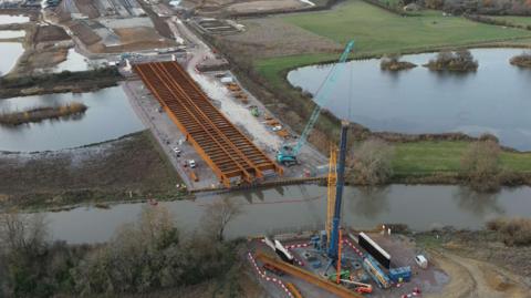 An aerial view of a viaduct skeleton made of large orange beams stretches from a road and across a river