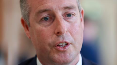 A close up photo of Paul Givan while he is speaking.   He has short, greying hair and is wearing a navy suit and a white shirt. 