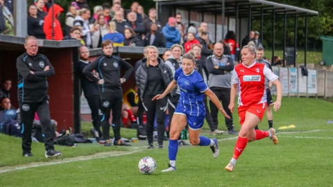 Two women on a football pitch. One wearing blue, the other wearing red. The blue player has the ball at her feet. In the background, the crowd and staff in black look at the players.