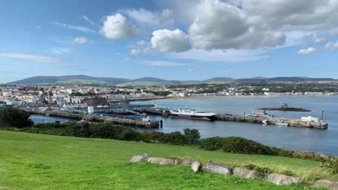 A view of Douglas on the Isle of Man, you can see boats in the harbour, the sea is blue. The monument the Tower of Refuge - a grey structure sits in the bay. Rows of houses and buildings line the seafront. There is green grass in the foreground and rolling hills in the distance. The sky is blue with white fluffy clouds.