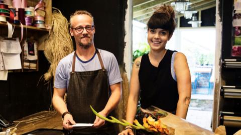 A man and a woman in overalls, standing in a florist shop, smiling at the camera.