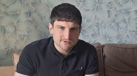 A young man looks at the camera. He has relatively short, dark hair and is wearing a dark polo shirt.