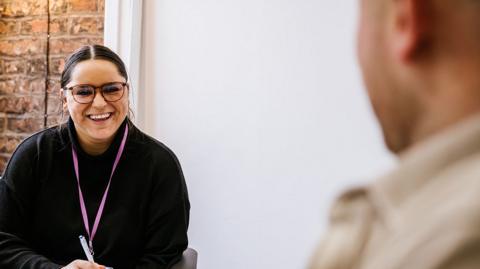 A staff member at the rehabilitation site with brown hair and brown glasses wearing a black top sits in front of a desk during a session with man who has his back to the camera and is blurred out.