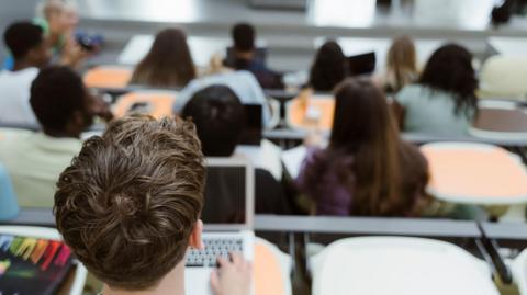 A group of students sits in a tiered lecture hall, listening to a presentation at the front of the room. In the foreground, one male student with brown hair types on a laptop.