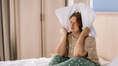 A woman sat on her bed in her pyjamas with a pillow wrapped around her ears. She is grimacing, as if being disturbed by noise.
