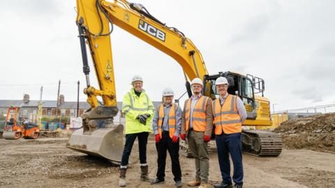 (L-R) Alex Isted, regional director at Morgan Sindall, Paul Latcham, leisure operations manager at Durham County Council, Cllr Joe Quinn, Cabinet member for resources, investment and assets, and Ben Kelsey, senior manager for projects and performance at Durham County Council. They are wearing hi-vis jackets  and hard hats and are standing in front of a large, yellow, JCB excavator.