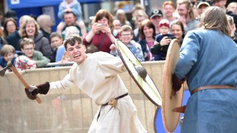Two re-enactors holding weapons and wearing replica medieval clothing face off against one another in a makeshift arena watched by a crowd of spectators. On the left, a young man wearing a white tunic looks angry as he prepares to swing an axe while holding a white and green, round shield. Facing him is another man wearing a blue tunic and holding a similar shield, whose back is to the camera. In the background, spectators are smiling and taking pictures.