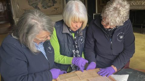 Three elderly women wearing Wentworth Woodhouse uniforms are examining an old document on yellowed paper. One woman points to it in a purple glove.