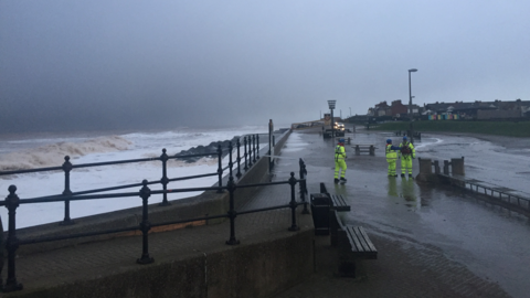 Sea wall and metal rails and rough sea on one side and three people in fluorescent stood on a wet promenade.