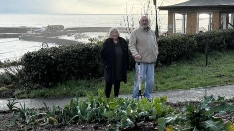 Cheryl and Alan Reynolds standing on a path at the community gardens in Lyme Regis with the harbour in the background.