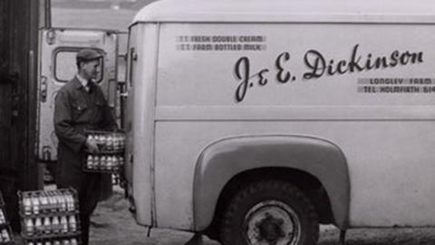 A black and white photo of a van reading 'J&E Dickinson'. A man is loading bottles of milk into the back of it