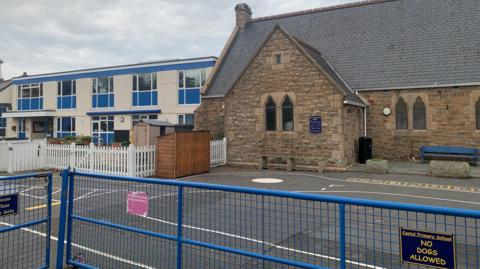 The outside of a school which is made up of two buildings - the right side one is brown and the left one is white and blue. There is a low blue gate surrounding it, with a sign saying Castel Primary School - no dogs allowed attached to the right side of it. 