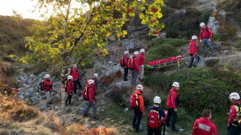 Mountain rescue team carrying a stretcher along a rocky hillside during a training exercise.