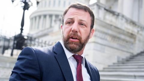 Markwayne Mullin wears a navy blue suit and a red patterned tie. In the background are white steps and the Capitol building in the distance. 