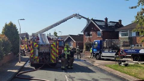 A fire engine can be seen with its aerial ladder platform lifting out of the top and reaching over to the roof of a house, which has been severely damaged by a fire. It is a sunny day and about seven firefighters can be seen outside the house, and a ladder is leant up to a window at the damaged house