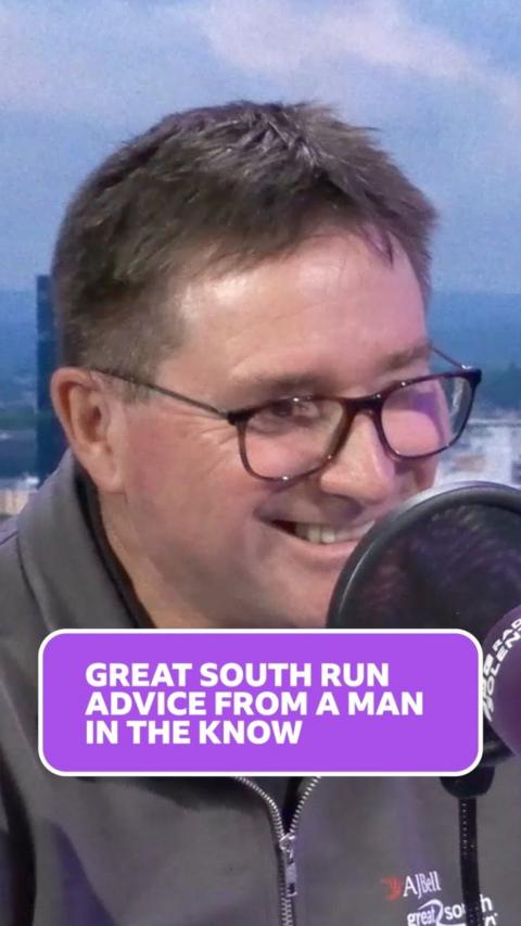 Dave Olsen smiles while in the radio studio, he has short light brown greying hair and tortoiseshell framed glasses