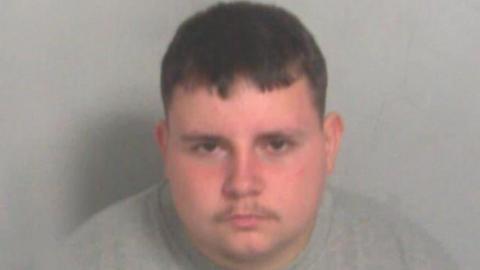 A custody photo of a man wearing a grey jumper. He is looking at the camera and has short brown hair.