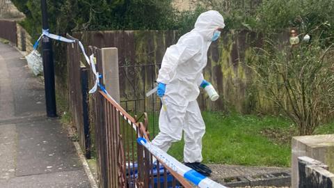 A person in full PPE white forensics clothing holding a white pot and a piece of plastic walking into a garden. There is police tape around the garden railings.