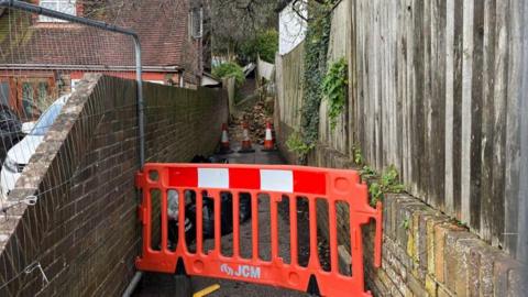 The narrow path with walls on either side which is blocked by a barrier and cones and further down a pile of what looks like rubble blocking the way.