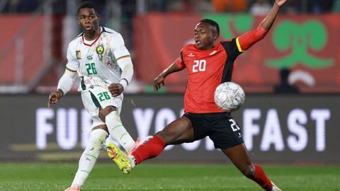 Footballer Christian Kofane, in a white kit with green, yellow and red detail and the number 26 in green on his chest and thigh, shoots to score for the Cameroon national team against Mozambique at the Afcon 2025. Mozambique player Oliver Keman, in a red shirt with black detail and black shorts, both with his number 20 on them in white, lunges across Kofane's path with his outstretched right leg as he attempts to intercept the ball, but it can be seen well past him
