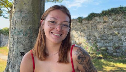 Lydia has ginger hair just passed her shoulders. She is wearing round, wire-rimmed glasses and has a nose ring in her right nostril. She's smiling at the camera and wearing a top with red straps on both shoulders. On her left shoulder is a tattoo stretching down her arm.