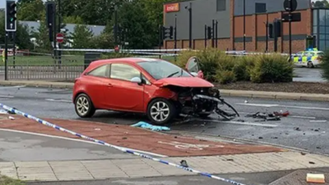 A stationary red car surrounded by police tape. The car has been involved in a collision and has sustained heavy damage to the front. Debris litters the road around the vehicle