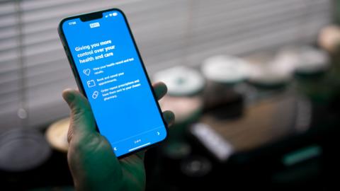 A smartphone is held up with an NHS menu visible on the screen.