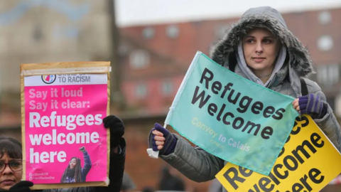 A woman standing outside holding a "refugees welcome" sign. She is wearing a hooded coat.