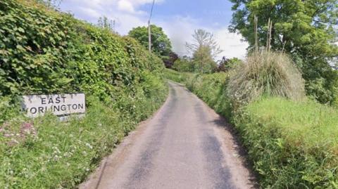 A sign reads EAST WORLINGTON and it is in a hedge next to a single track road. The hedge is lush and has pink and white wild flowers. It is sunny.