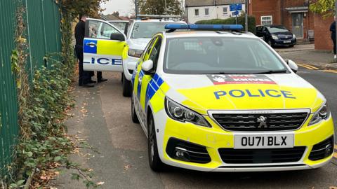 A white, yellow and blue police car stands on a road in front of a white, yellow and blue police truck that has its front right door open. Two uniformed police officers stand near the open door of the truck. There is a set of green park railings to the left of the police cars. Some houses stand to the right-rear of the photo.