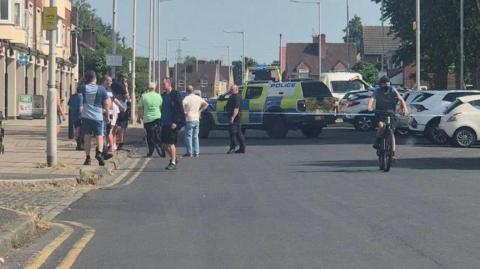 A group of local residents stand in front of a police cordon by a row of terraced townhouse type buildings with shops on the ground floor. Police vehicles and an officer in black uniform are visible behind the cordon. 