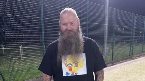 Alex Hammond smiles at the camera on a football pitch. He has a long beard that is down to his chest. He has grey hair that has been slicked back. He wears a black T-shirt with an image of a yellow teapot on it. 