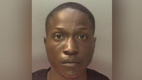 Police mugshot of a man with close-shaven black hair and wearing a black T-shirt. He is looking straight at the camera.