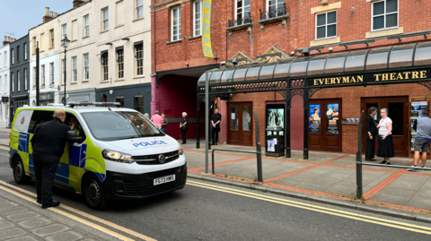 A police van parked with an officer learning into the driver's side, against the backdrop of a theatre