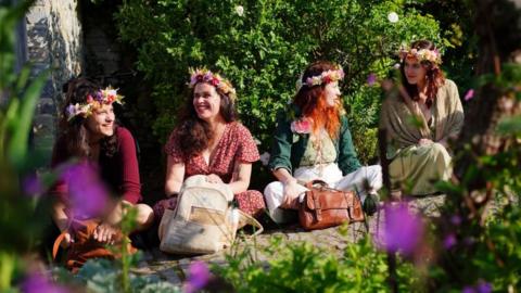 Four women sitting on the ground in the sun. They are wearing flower crowns and smiling