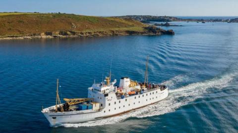 A white ferry sails out to sea, the Isles of Scilly behind