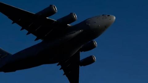 US military plane flies overhead, it is dark grey against a blue sky
