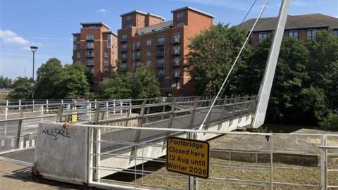 IMage of the footbridge which has a yellow sign in the foreground which reads FOOTBRIDGE CLOSED HERE 12 AUG UNTIL WINTER 2026
