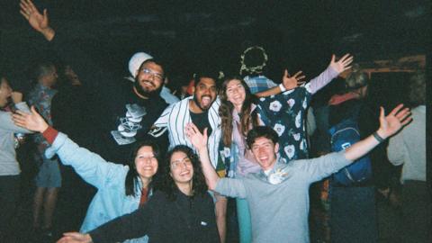 A group of young people. Some are standing and some are kneeling and they all have their arms outstretched. They are all smiling widely at the camera. There are three young women and three young men and they appear to be at a festival. It is dark behind them and other people can be seen in the background.