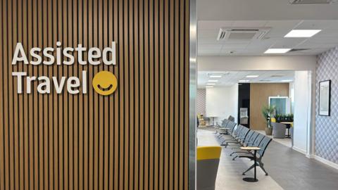 A photo taken from the entrance to the assistance lounge. On the left is the reception area with the words Assisted Travel in white on the wooden panelled wall. Beyond it to the right is the lounge itself in muted greys, whites and yellows. There are large frames on the wall awaiting artwork, rows of widely spaced chairs, plants and tables.