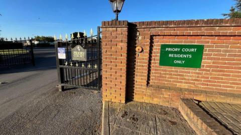 The gates of Priory Court Caravan Park. There is a brick wall connecting to an iron fence. 