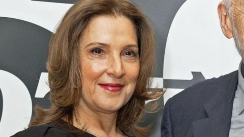 Barbara Broccoli at an event celebrating 60 years of James Bond, standing in front of a "007" backdrop. She is dressed in black.