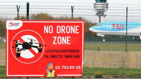 A large red sign at Brussels Airport reads “NO DRONE ZONE” in white letters, with a black drone crossed out inside a red circle. A cartoon airport worker wearing a yellow safety vest is shown pointing to the message. Behind the fence, a blue and white TUI airplane on the runway.
